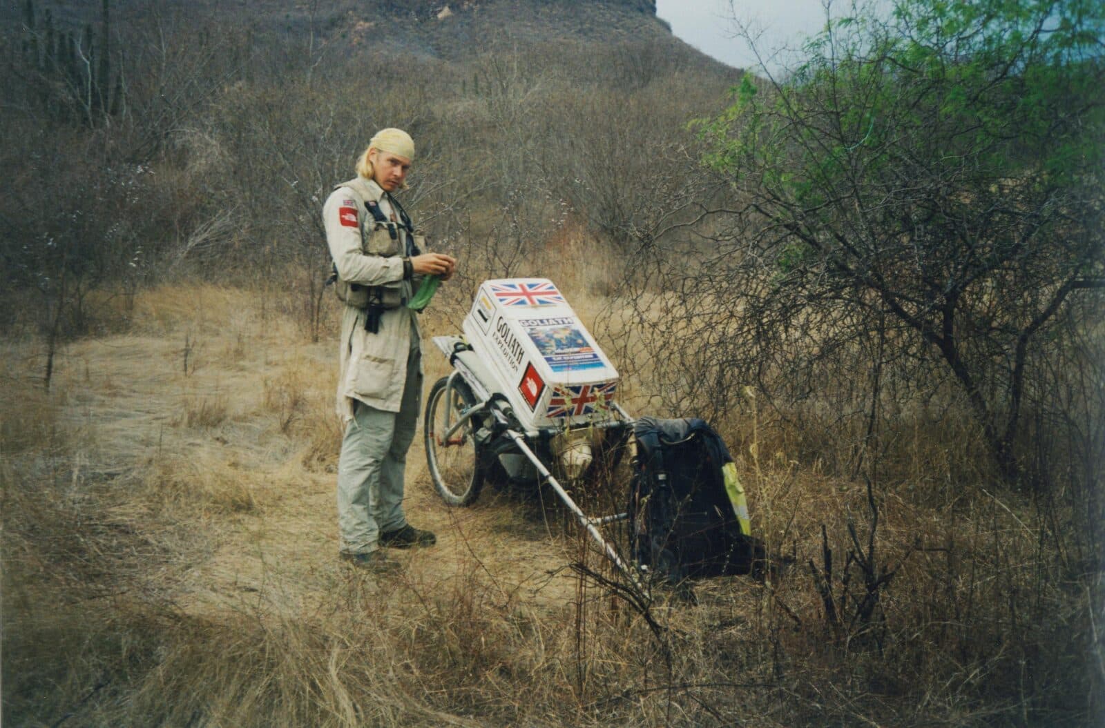 Karl Bushby with his Goliath Expedition cart on arid scrubland in Mexico, dry hills rising behind him.