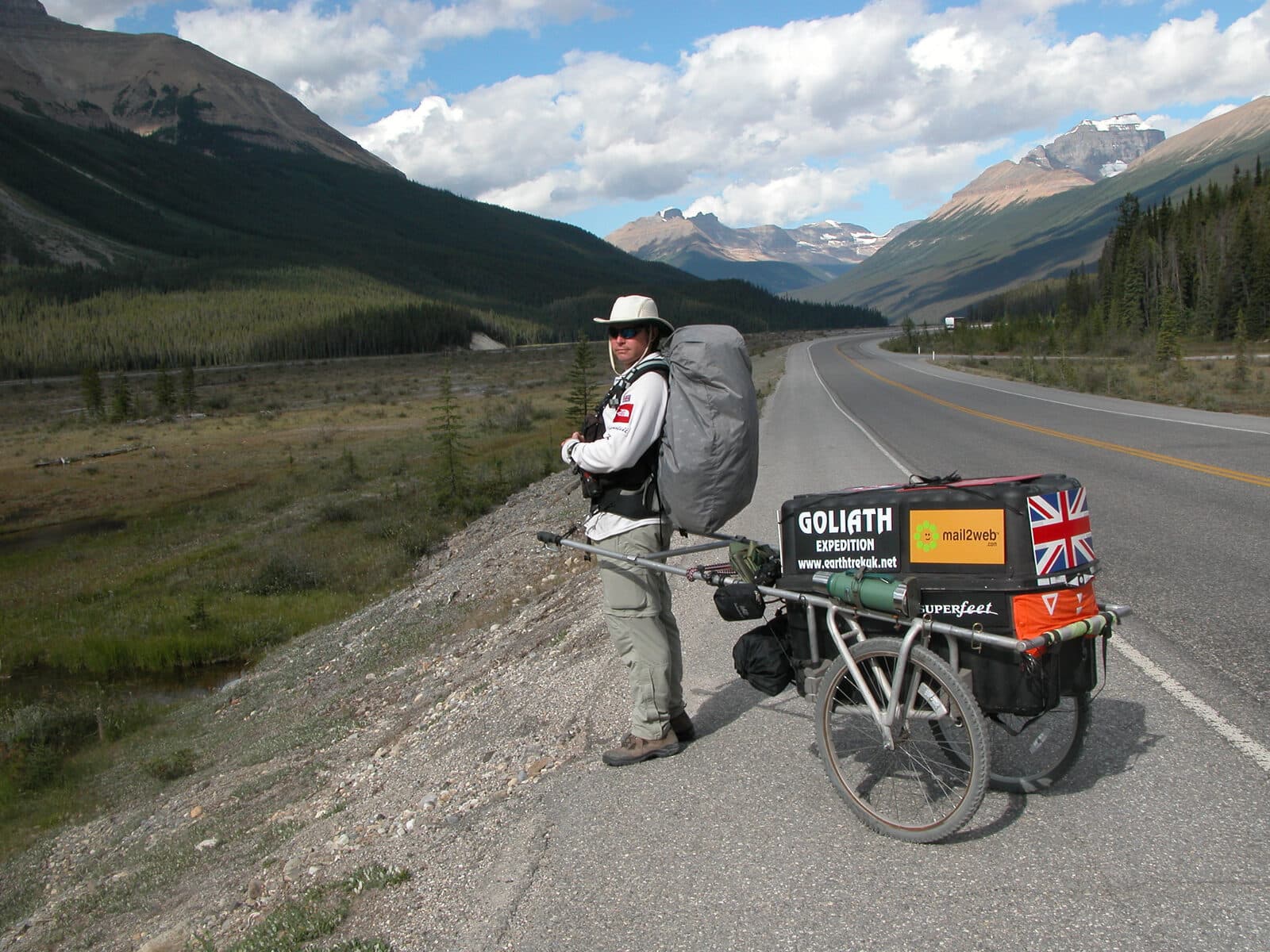Karl Bushby hauling his cart along an empty highway through the Canadian Rockies, snow-capped peaks ahead.