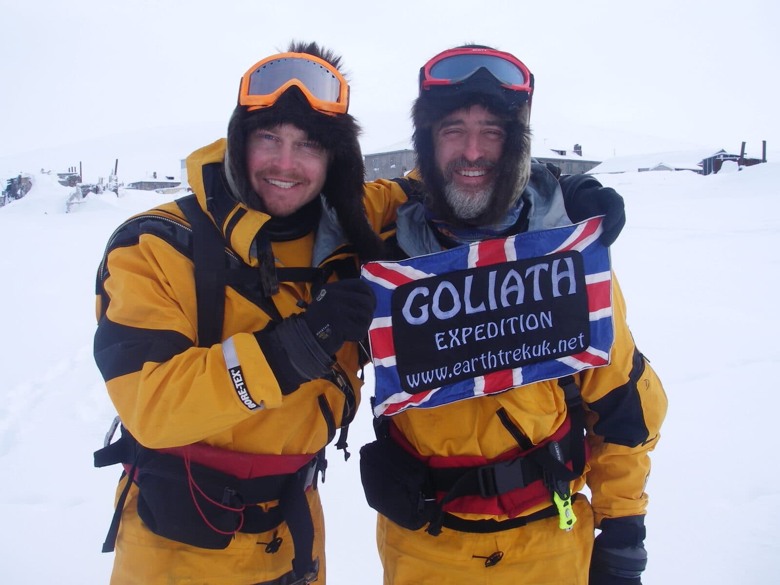 Karl Bushby and his crossing partner in fur hats and survival suits, holding the Goliath Expedition flag after reaching Uelen, Russia.