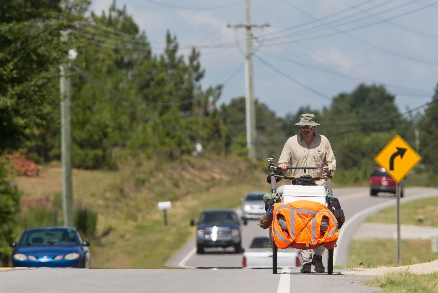 Karl Bushby pushing his cart along a roadside in the United States, traffic passing on a summer highway.