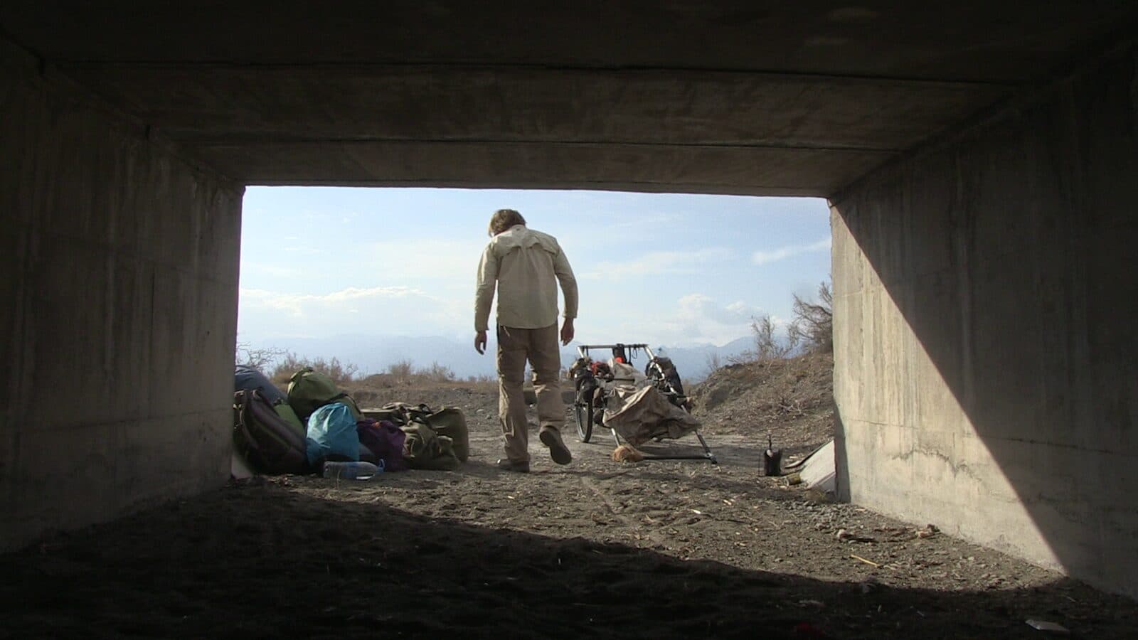 Karl Bushby walking out of a concrete underpass toward distant mountains in China, his cart and kit beside him.