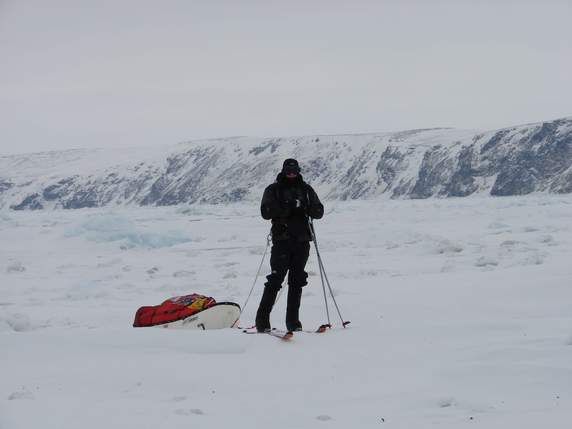 Karl Bushby on skis hauling a red sled across the frozen Russian Arctic, snow-covered cliffs behind him.